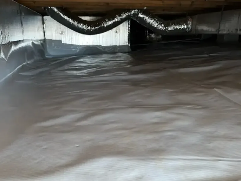 A crawl space under a raised floor with a white vapor barrier and flexible ducts